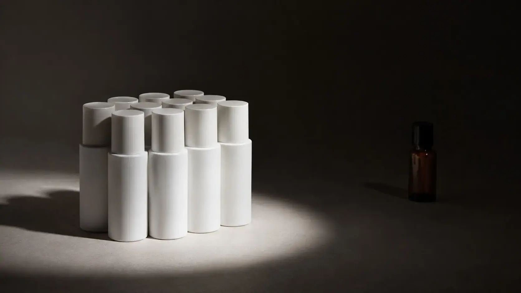 Cluster of white cosmetic bottles standing in a spotlight, one small amber bottle set apart in shadow